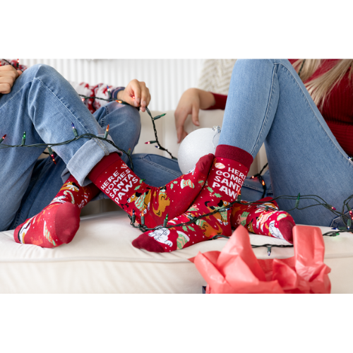 Two people wearing festive red socks with Christmas-themed text, sitting on a couch with holiday lights and presents.