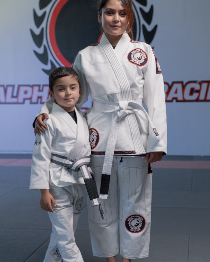 Two individuals in white martial arts uniforms standing in a dojo with a logo in the background.