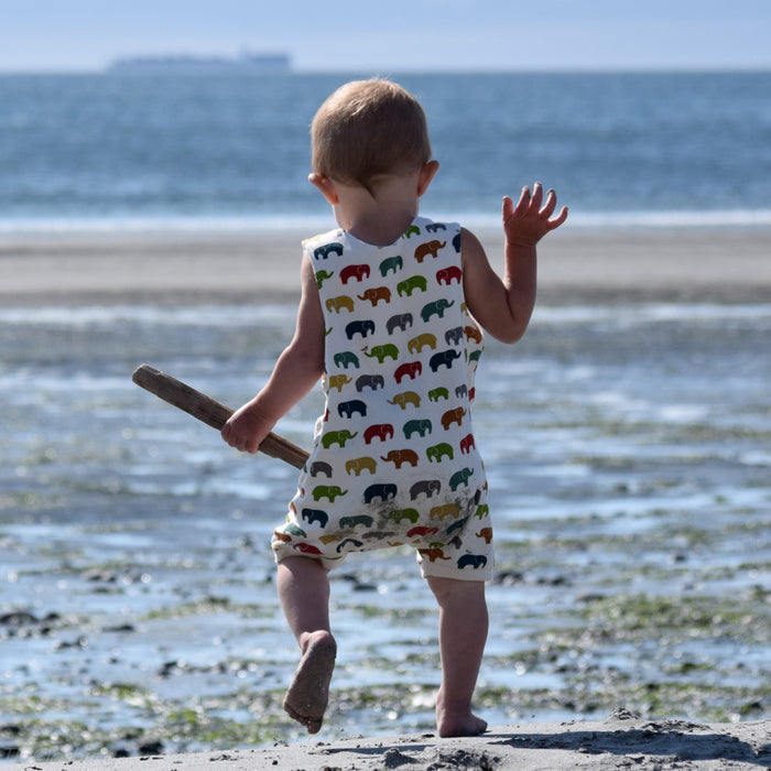 Toddler walking away on a sandy beach holding a piece of driftwood, wearing a sleeveless organic cotton romper with a colorful elephant print made by TK Clothing